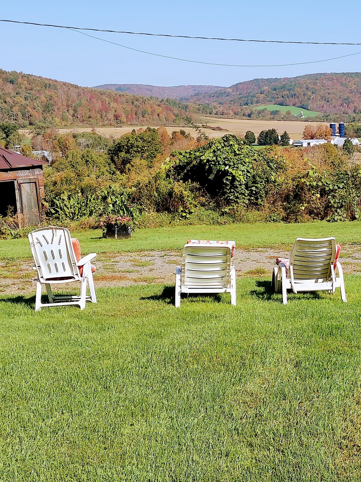 Three white lounge chairs are positioned on a grassy area, facing a wide view of rolling hills and fields. Autumn foliage in warm tones colors the landscape, with a clear sky overhead, creating a serene outdoor space for relaxation.