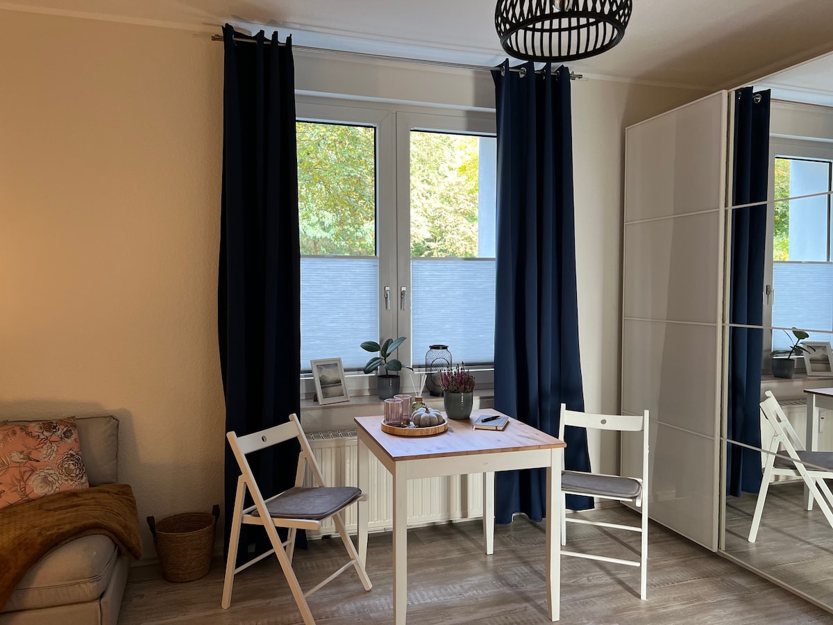 A bright corner of the apartment features a small wooden dining table flanked by two white chairs. A cozy sofa is partially visible, and large windows provide natural light, adorned with dark blue curtains. A mirrored wardrobe adds depth to the space.