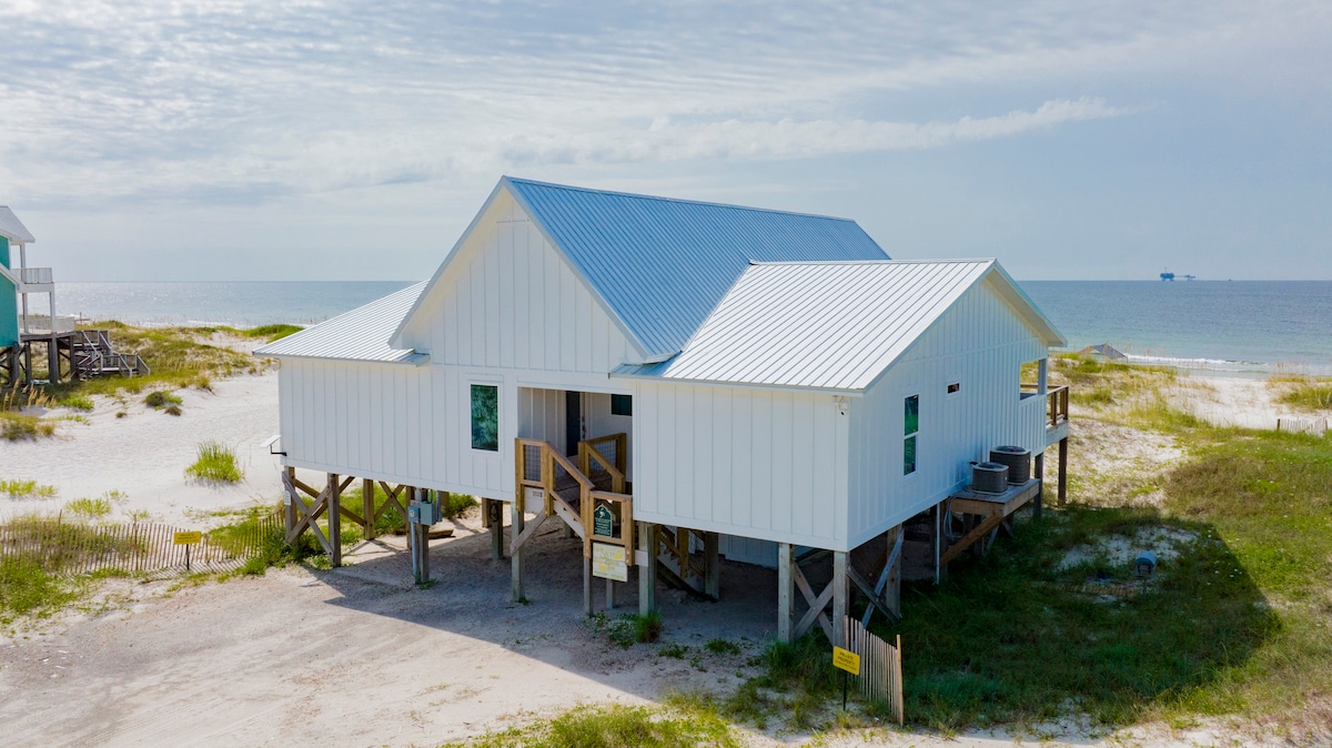 A contemporary white beach house is shown from an elevated angle, highlighting its elevated structure on stilts. The ocean is visible in the background, while beach grasses surround the property, creating a natural transition between the home and the shoreline.
