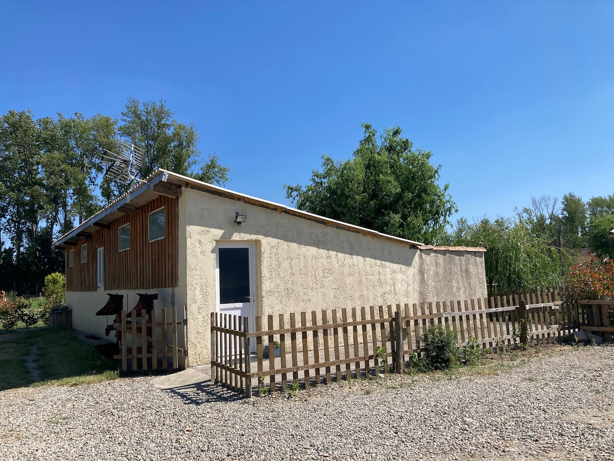 The exterior of the gîte is depicted, featuring a simple design with wooden accents. Large windows provide natural light, while a fenced area surrounds the property. Mature trees and greenery add to the serene setting, and a gravel driveway leads up to the entrance.