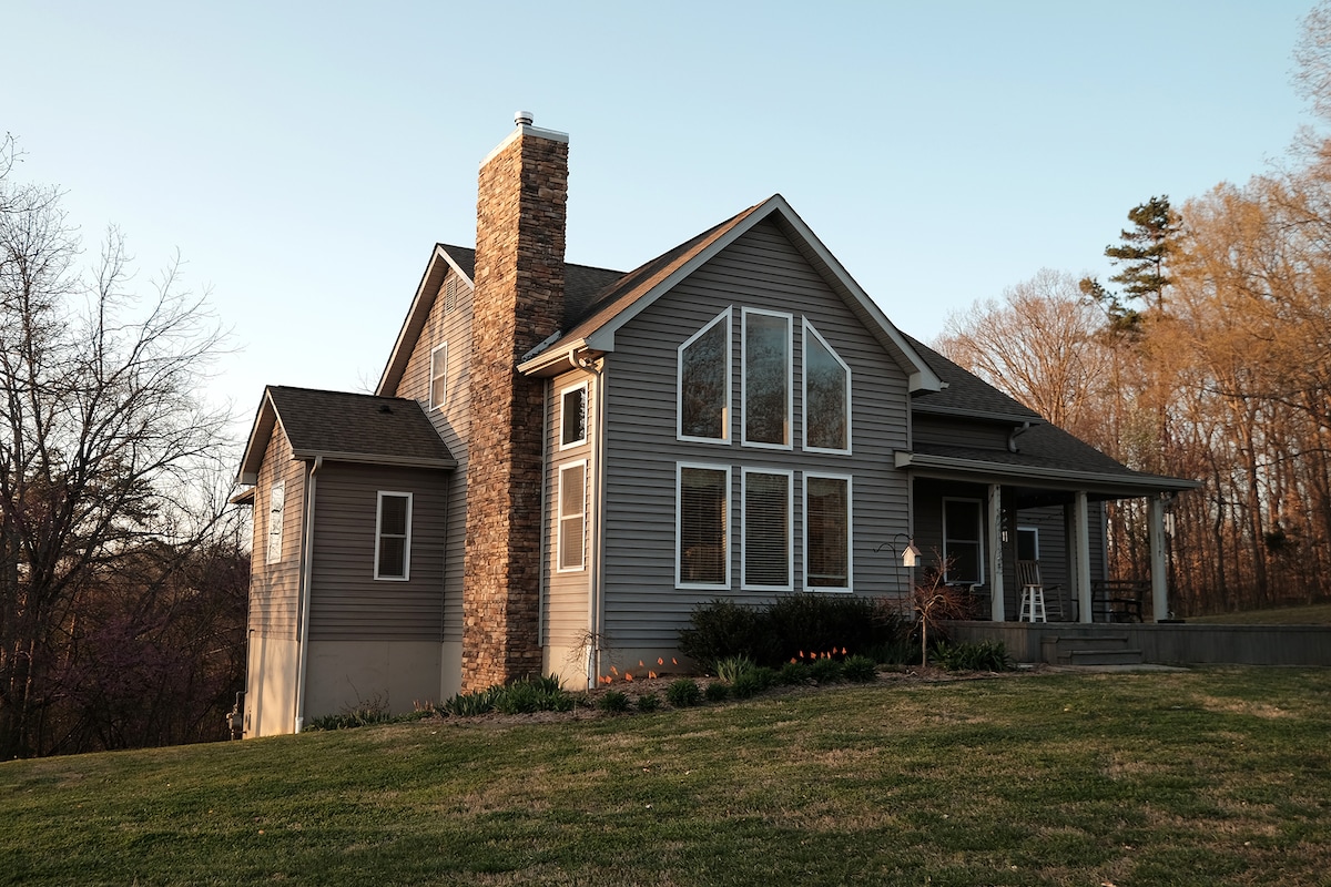 The home's exterior showcases a contemporary design, featuring a combination of stone and gray siding. Large windows allow natural light to enter, and a spacious porch extends towards a lush green lawn, which is complemented by seasonal flowers. Surrounding trees contribute to the serene setting.