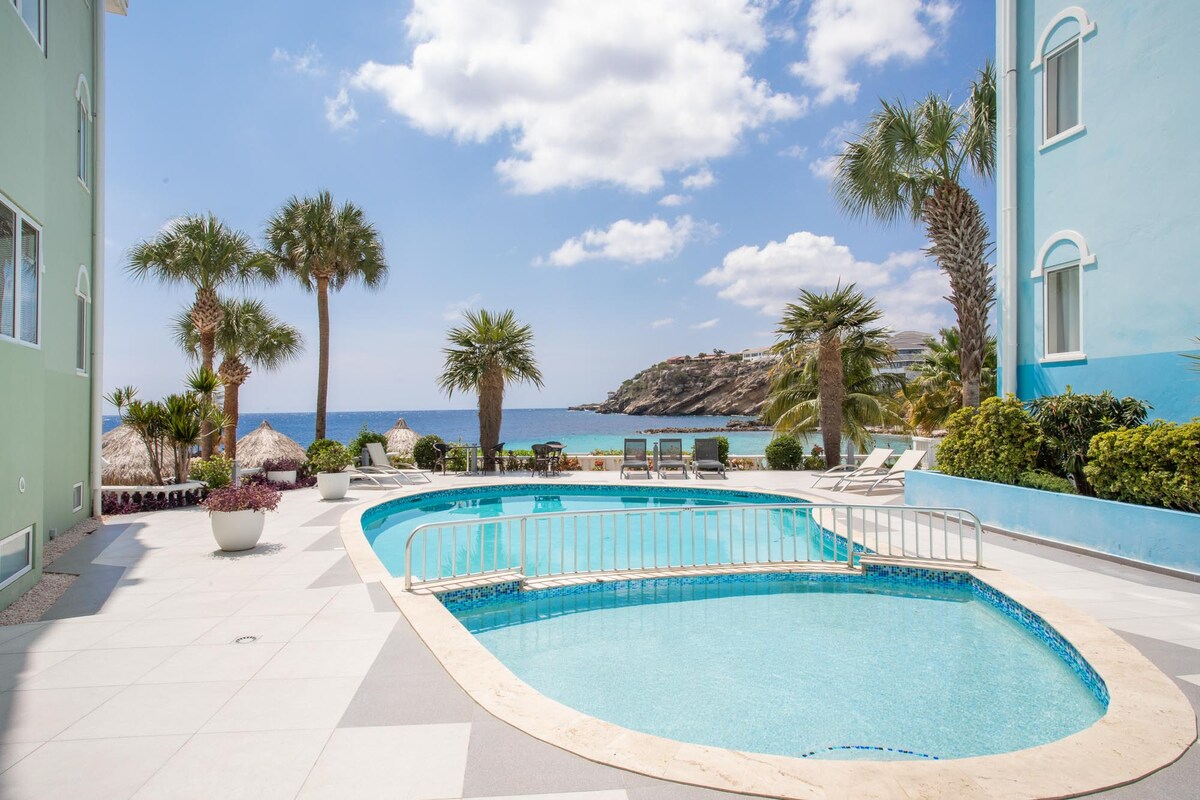 A serene outdoor area features a private swimming pool surrounded by lounge chairs, palm trees, and tropical plants. The clear blue water of the pool contrasts with the vibrant blue sky and distant shoreline, creating a relaxing atmosphere ideal for unwinding.