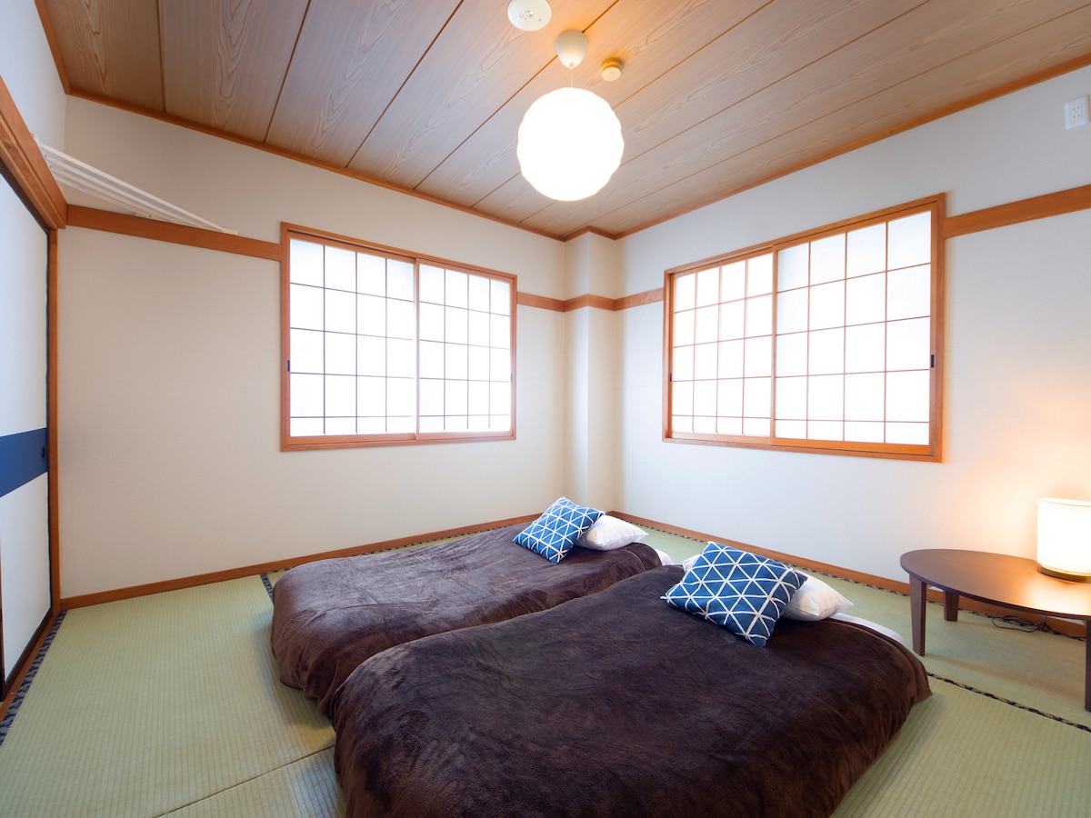 A serene Japanese-style room features tatami flooring and warm wooden walls. Two futons are placed side by side on the floor, adorned with blue patterned cushions. Natural light filters through two shoji-style windows, complemented by a simple wooden table and a soft glowing lamp.