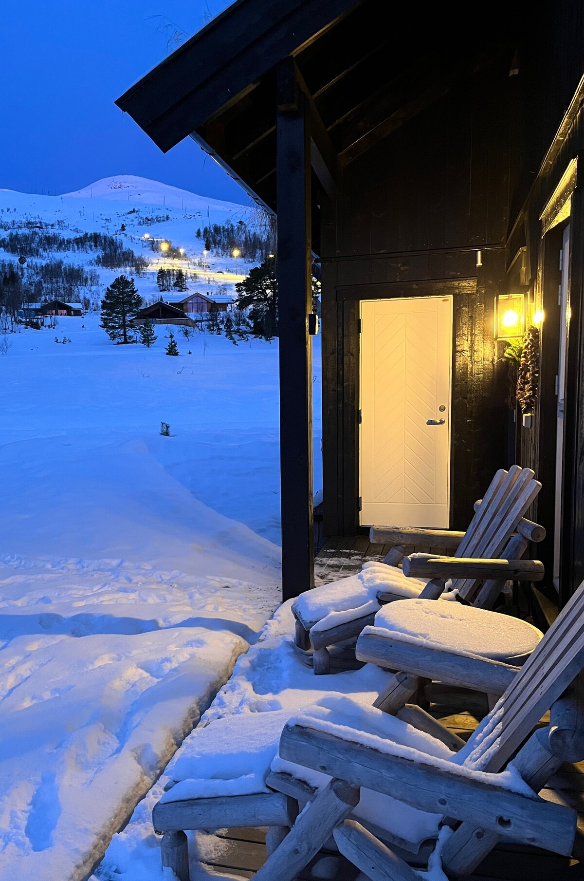 An entrance porch features two rustic wooden chairs, lightly covered with snow. A door painted in white stands prominently, illuminated by a warm light. The backdrop reveals a winter landscape with snow-covered hills and soft lights in the distance.