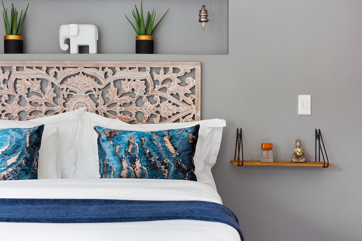 A serene sleeping area features a carved wooden headboard, complemented by decorative pillows with blue and gold patterns. Simple green plants are placed in black pots, while a small shelf displays a candle and figurine. Soft light is provided by hanging pendant lights.
