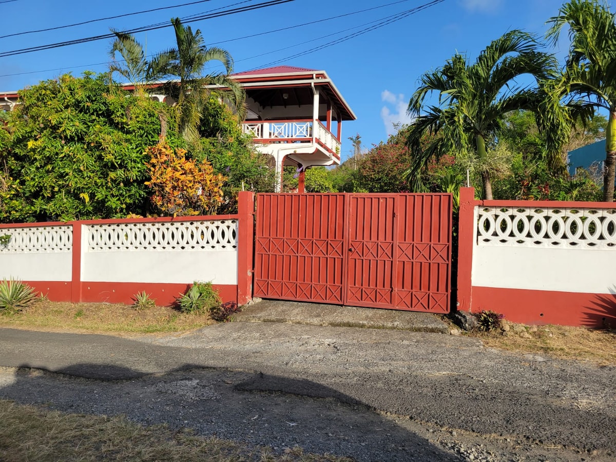 A vibrant red gate is set within a white and red boundary wall, surrounded by lush tropical foliage. The property is framed by palm trees and other greenery, contributing to a welcoming entrance. A clear blue sky is visible in the background.