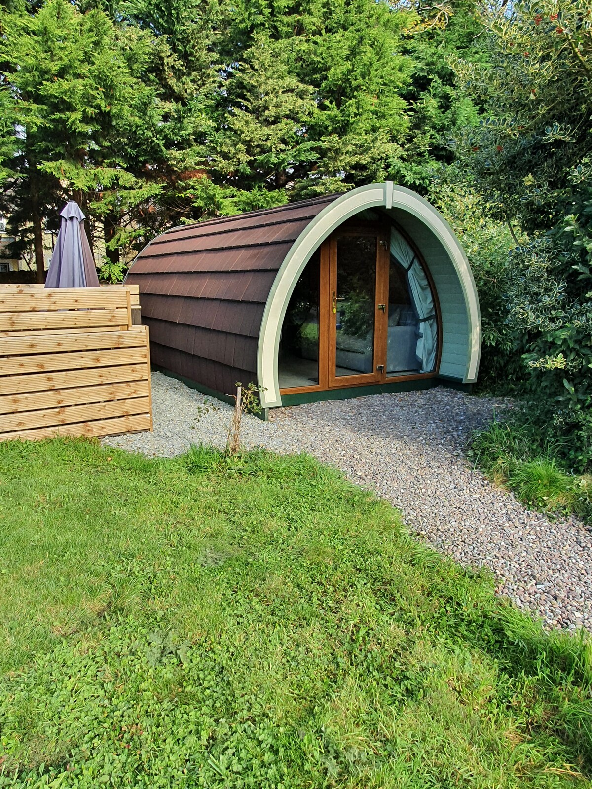 The glamping pod is nestled in a lush green area, showcasing a curved wooden exterior. Large glass doors provide a glimpse inside, framed by natural foliage. A gravel pathway leads to the entrance, enhancing the connection to the surrounding landscape.