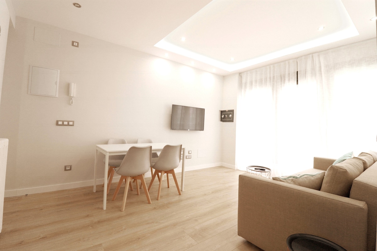 A well-lit living and dining area is shown, featuring a modern white dining table with four chairs. A large window allows natural light to fill the space, complemented by light-colored hardwood flooring. A comfortable sofa is positioned nearby, with a wall-mounted television visible.