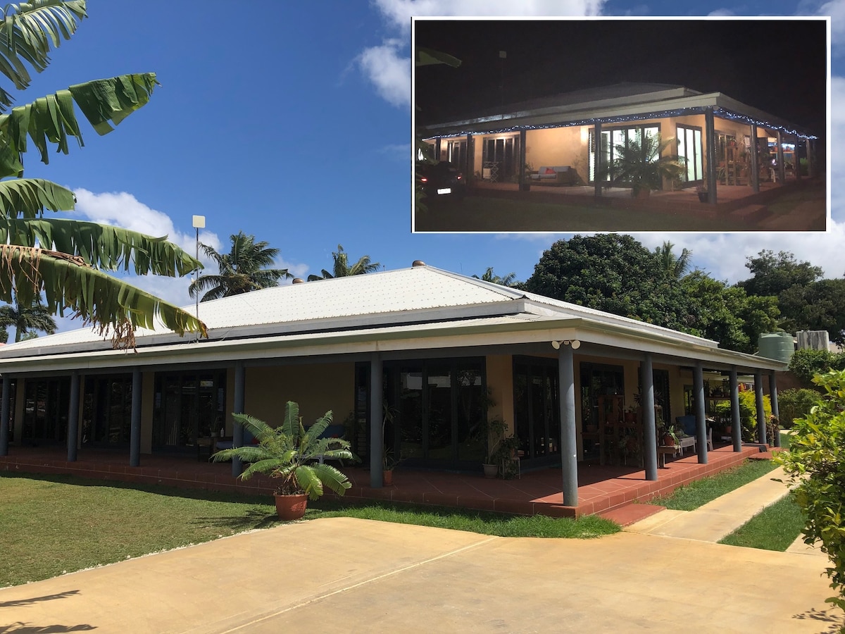 The exterior of a spacious Bed & Breakfast is shown, featuring a modern design with large windows and a wrap-around veranda. Lush plants are placed around the property, with a well-maintained lawn giving a welcoming appearance. The main entrance is easily accessible from the driveway.