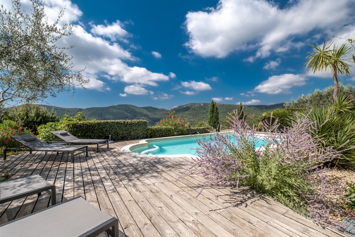 A serene outdoor space features a rectangular pool surrounded by wooden decking. Lounge chairs offer comfort, while vibrant greenery, including flowering plants and palm trees, frames the scene. The distant hills provide a scenic backdrop under a partly cloudy sky.