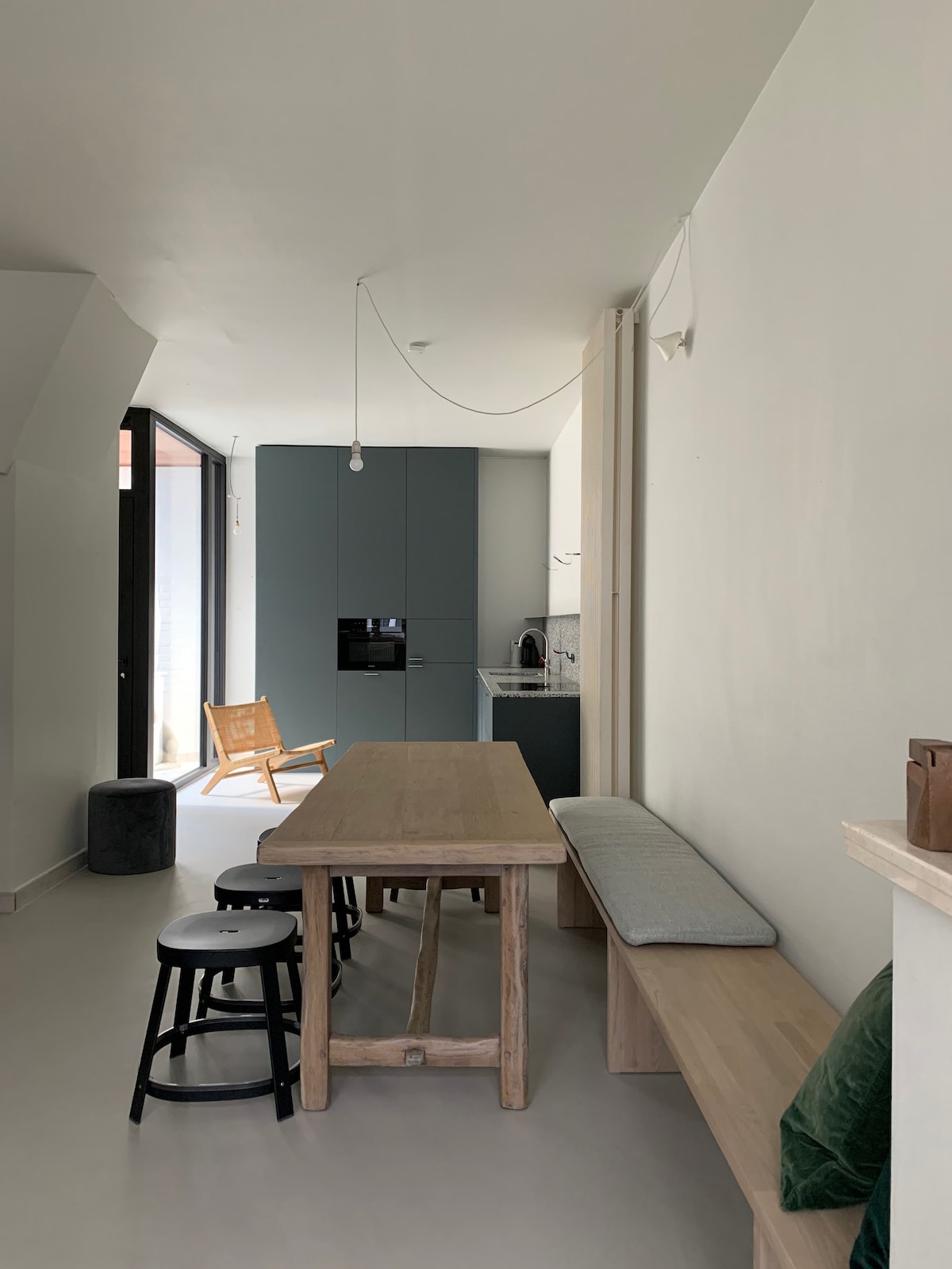 An open kitchen and dining area is presented with a wooden table and a bench along one side. Minimalist stools in black and white are positioned nearby. A modern kitchen with dark cabinetry is visible in the background, complemented by a cozy seating chair beside a large window.