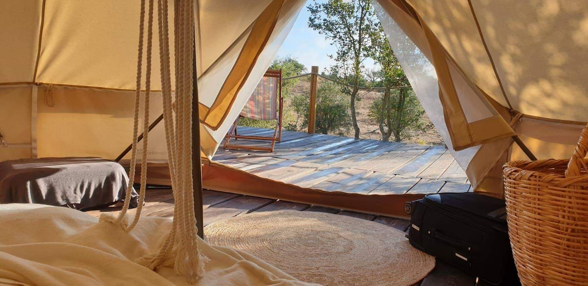 The image shows the entrance of a spacious tent, framed by soft fabric that gently drapes. Outside, a wooden deck is visible, surrounded by greenery. Natural light streams in, illuminating a cozy sleeping area with a rug and personal luggage. A welcoming atmosphere is suggested.