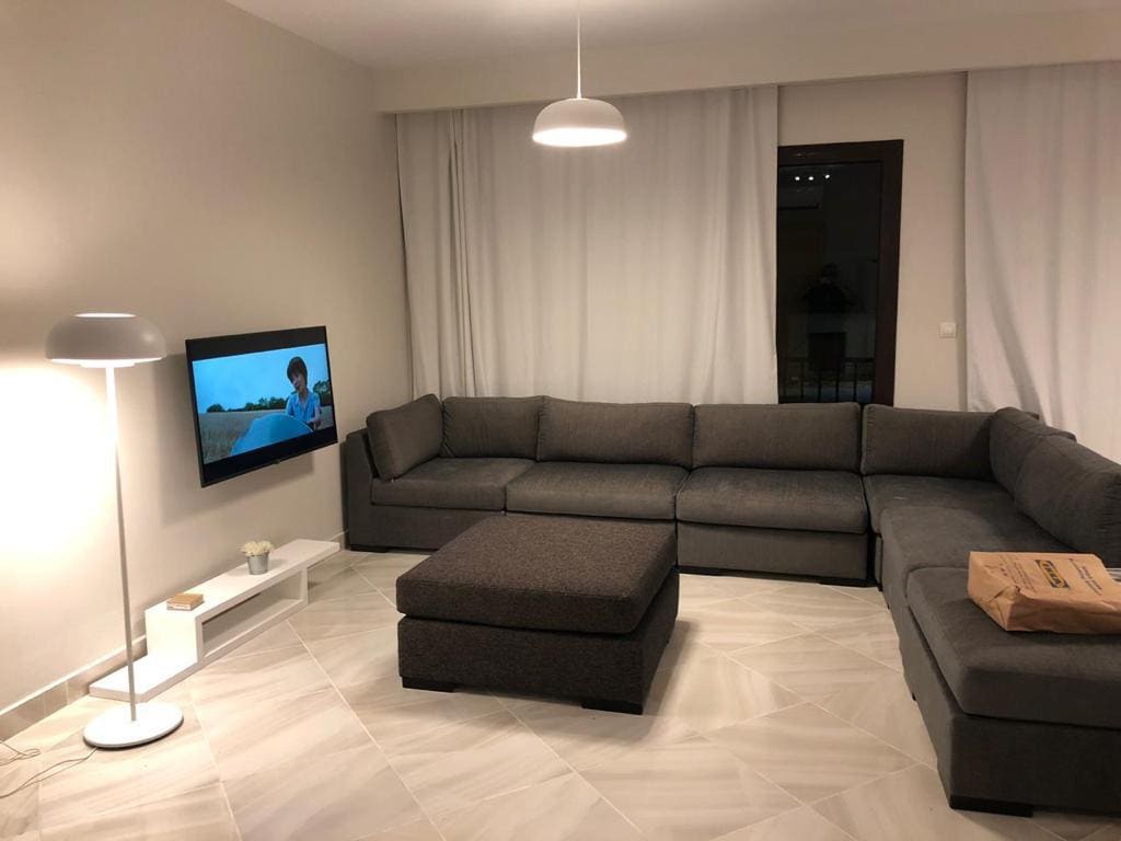 A spacious living room is featured, showcasing a large sectional sofa in a dark grey fabric. A minimalist white coffee table sits in front, and a flat-screen television is mounted on the wall. Light-colored curtains frame the window, allowing natural light to enhance the space.