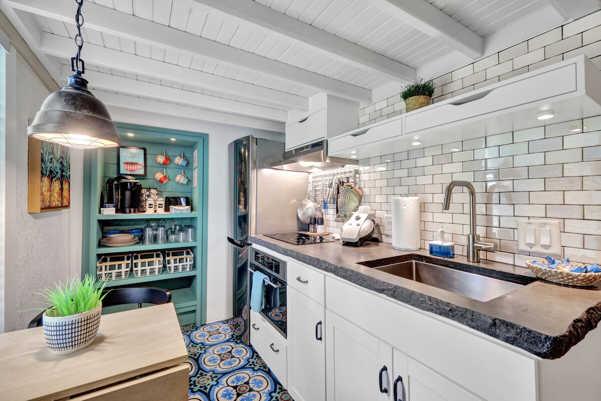A fully equipped kitchen is presented, featuring sleek white cabinetry and a black countertop. Modern appliances, including a refrigerator and oven, are visible. The tiled floor exhibits a vibrant pattern. Natural light enters from a nearby window, enhancing the functional layout.