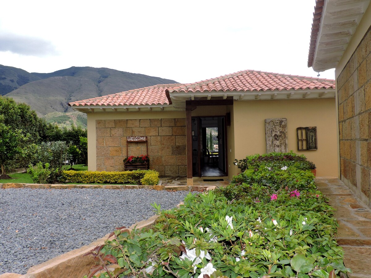 The exterior of the cabin features a welcoming entrance with a stone façade and a tiled roof. Lush greenery surrounds the pathways, enhancing the view of the mountains in the background. A sign reading 'WELCOMES' is prominently displayed, inviting guests into the cozy space.