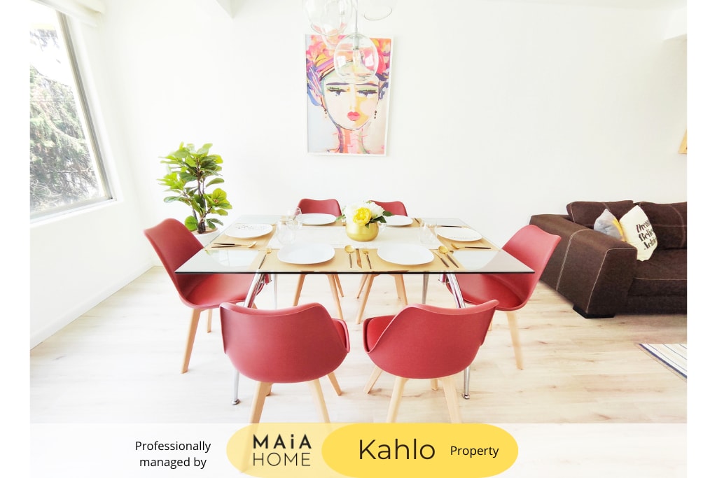 A modern dining area features a glass table surrounded by six red chairs, set with plates and utensils. Natural light floods in through large windows, illuminating a potted plant in the corner. A cozy sofa is visible in the background.