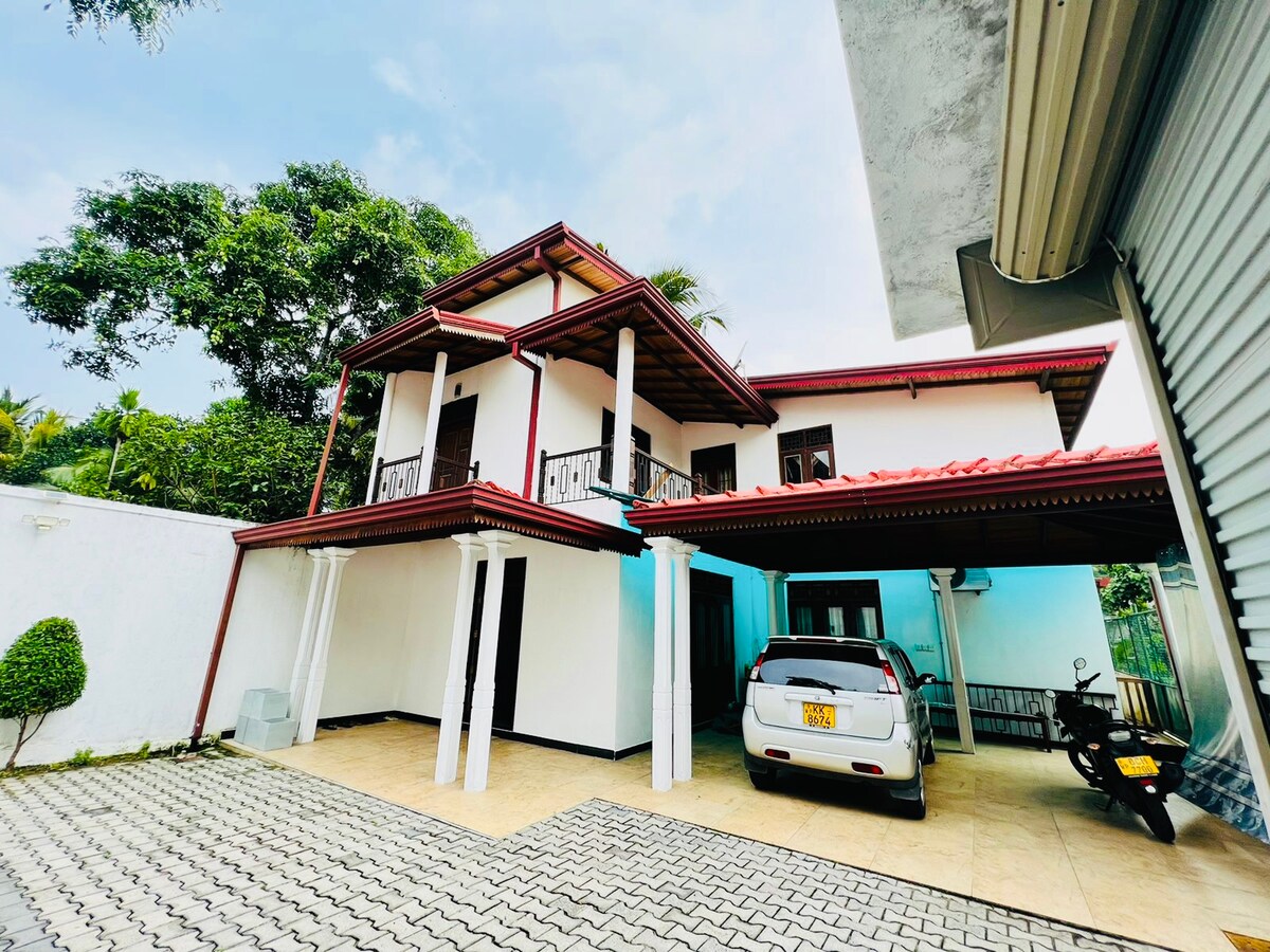 A two-story home is depicted, featuring a covered parking area that accommodates multiple vehicles. The exterior is characterized by white walls and a red roof, complemented by lush greenery surrounding the property. Balconies provide a view of the outdoor space.