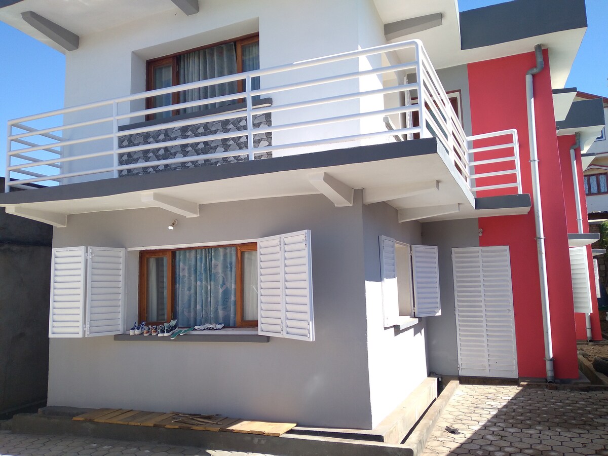 A modern two-story building features a combination of white and grey walls, with a bold red section on one side. Large windows with white shutters allow natural light to fill the spaces. A small balcony is positioned above, providing additional outdoor space.