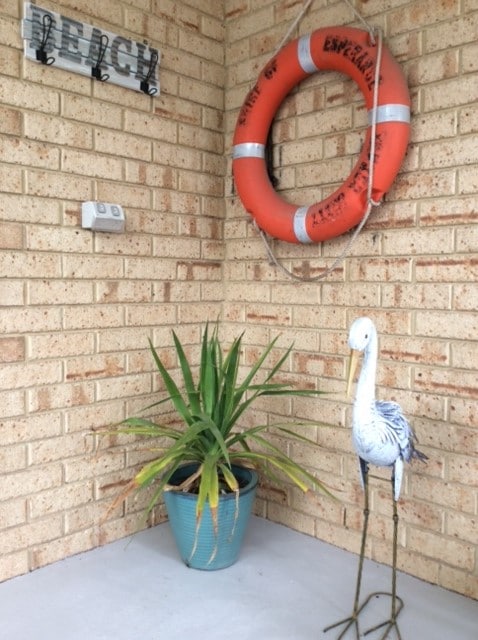 A welcoming entryway features a terracotta-colored life ring mounted on a brick wall. A potted plant adds greenery beside a decorative metal bird, creating a charming coastal vibe. The space is neatly arranged with wall hooks for convenience.