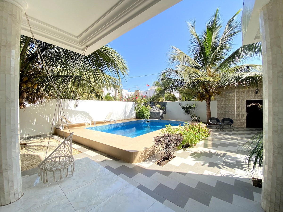 An outdoor space is highlighted by a large pool surrounded by palm trees and greenery. A shaded area features a hanging chair, while patterned tiles lead to the water, creating a relaxing atmosphere. Clear blue skies complete the inviting scene.