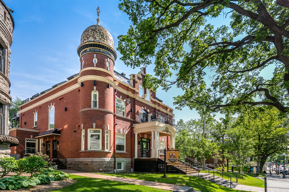 The exterior of a historic building is showcased, featuring red brick walls and ornate architectural details. A dome crowns the structure, while large windows allow for natural light. Surrounding trees and greenery enhance the inviting atmosphere of the property.