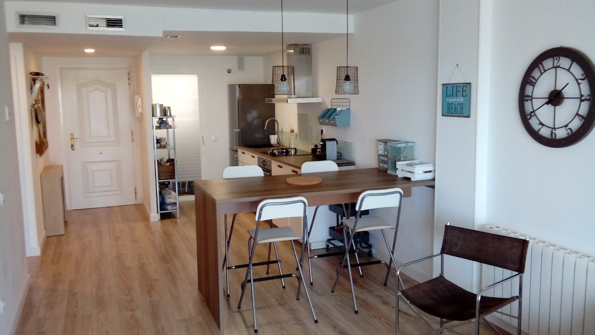 A modern kitchen area features a spacious wooden dining table with sleek, white chairs. The open layout includes a well-equipped kitchen with stainless steel appliances. A wall clock and decorative signs add subtle charm to the light-filled space.