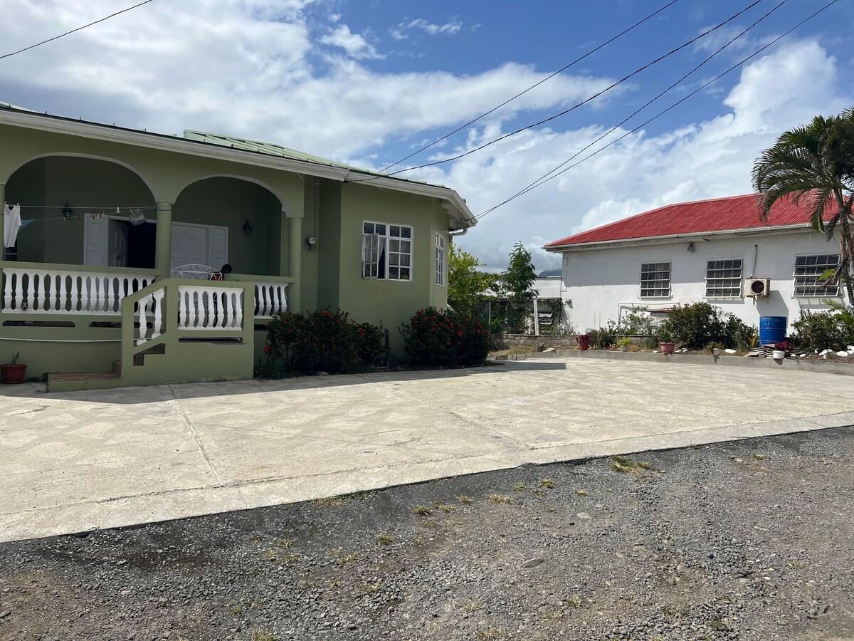 A modest house is shown with a welcoming front porch and decorative railing. Lush greenery lines the entrance, while a nearby structure with a red roof complements the scene. The spacious driveway provides ample parking space, framed by gravel and paved areas.