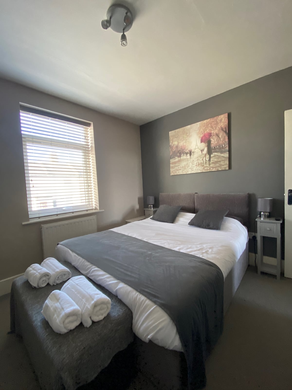 A well-appointed bedroom features a spacious bed with neutral bedding, accented by a dark throw. A piece of wall art depicting a serene scene enhances the room. Natural light filters through a window with blinds, creating a calm ambiance. Fresh towels are neatly arranged on the foot of the bed.