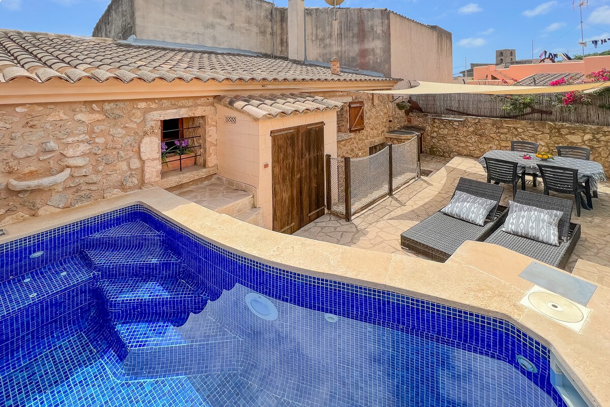 An outdoor area features a spacious jacuzzi-integrated pool, surrounded by a stone terrace. A dining table with chairs and lounge seating is visible, complemented by vibrant bougainvillea climbing nearby walls. The rustic charm of the home is highlighted by natural stone and a tiled roof.