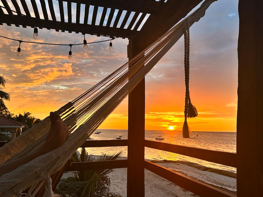 A hammock draped between wooden posts offers a view of the sunset over the ocean. The sky is filled with oranges and yellows as boats float on the calm water. Warm lighting from string lights enhances the serene atmosphere.