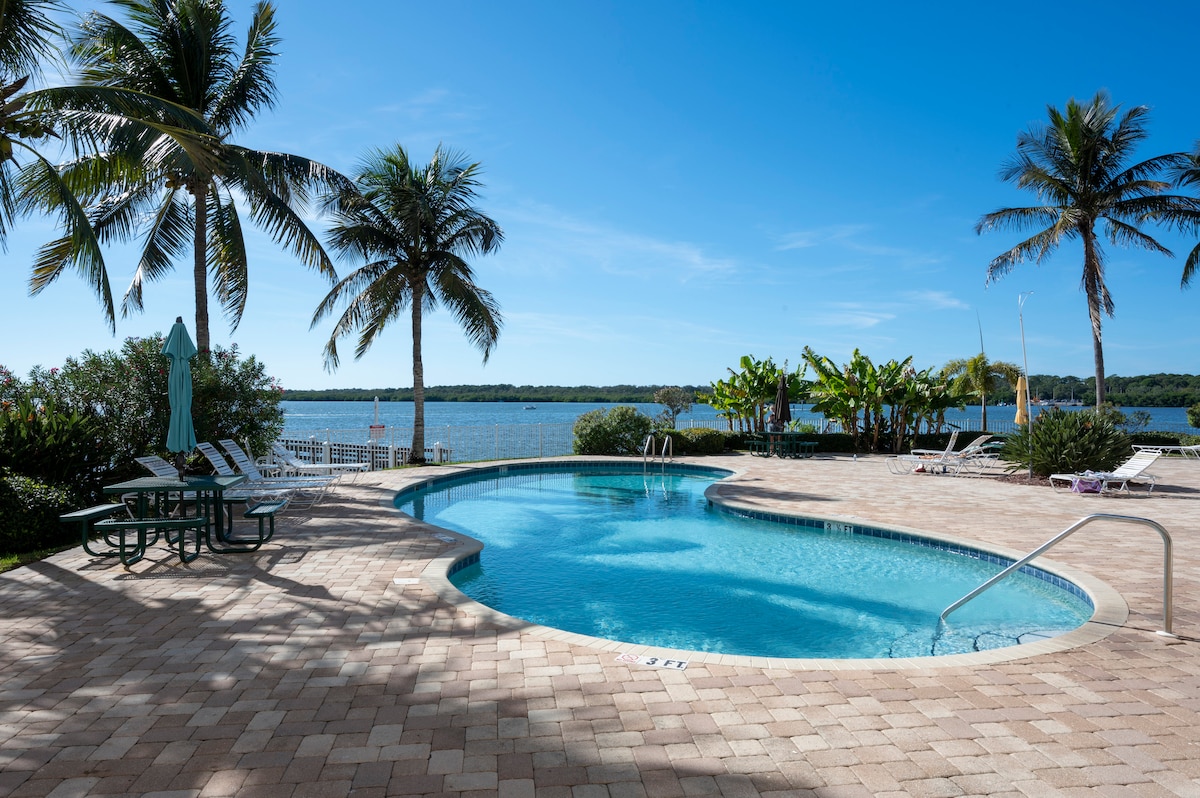 A serene pool area is positioned beside the water, surrounded by palm trees. Lounge chairs are available on the patio, offering views of the bay. A dining table with chairs is placed under a shaded umbrella, providing a relaxing outdoor space.