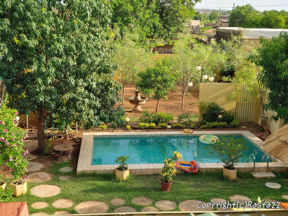 A refreshing outdoor space features a clear pool surrounded by lush greenery and vibrant plants. A small area with stepping stones leads to the pool, while various seating options and a fountain contribute to the tranquil setting.