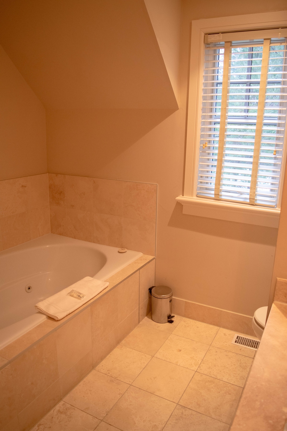 A serene bathroom features a spacious jacuzzi-style tub alongside beige tiled walls and floor. A small trash bin is positioned next to the tub. Natural light filters through a window with wooden blinds, creating a calm environment.
