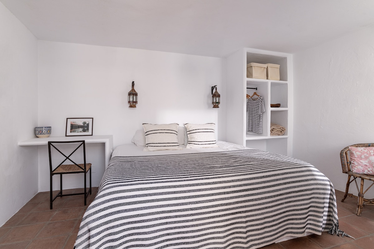 A bright room features a spacious bed with a patterned blanket, accompanied by wall-mounted lanterns. A small desk with a chair is positioned against one wall, while shelving displays folded clothing and a framed picture, enhancing the minimalist design.