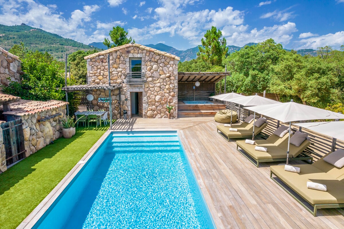A stone house sits amidst greenery, featuring a shimmering pool surrounded by sun loungers under large umbrellas. The deck showcases a blend of wood and grass, providing a spacious area for relaxation, with scenic mountain views in the background.