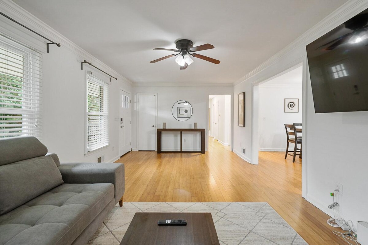 A living area features hardwood flooring and a comfortable gray sofa. A round mirror is mounted above a console table, and a ceiling fan provides ventilation. Light filters through window blinds, while a flat-screen TV is mounted on the wall, adding to the inviting atmosphere.