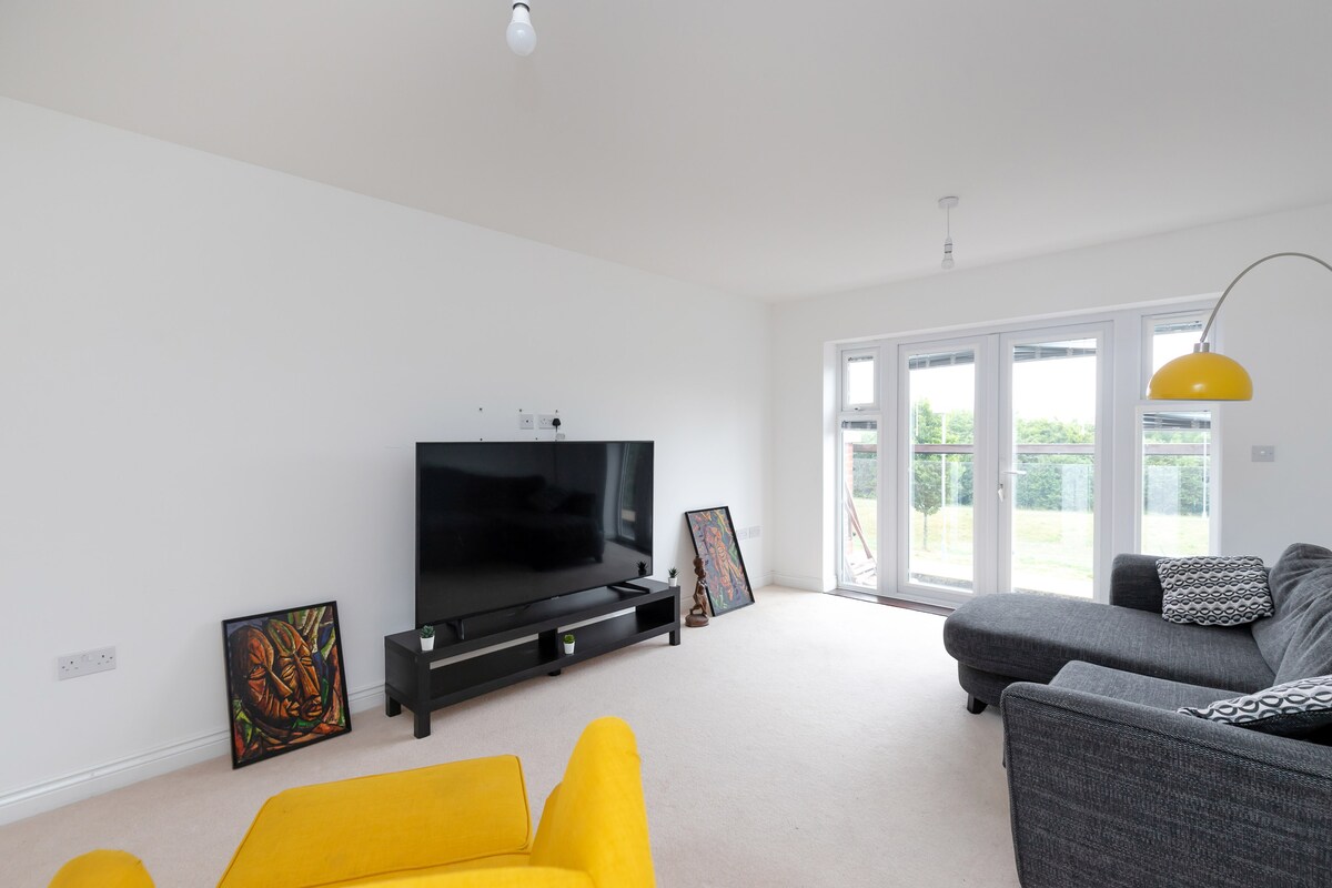 A bright living area features two large windows that provide views of the outdoor space. A modern television is positioned on a black console table, and a gray sectional sofa sits adjacent. Two colorful artworks add visual interest to the neutral-toned walls.