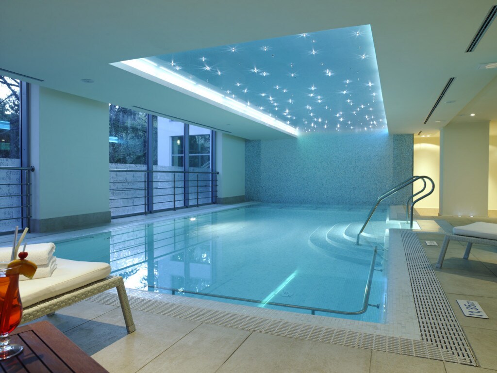 The indoor pool area is illuminated by soft, star-like lights on the ceiling. The pool features clear water, a gentle entry with steps, and is surrounded by sleek tiles and large windows that allow natural light to fill the space.