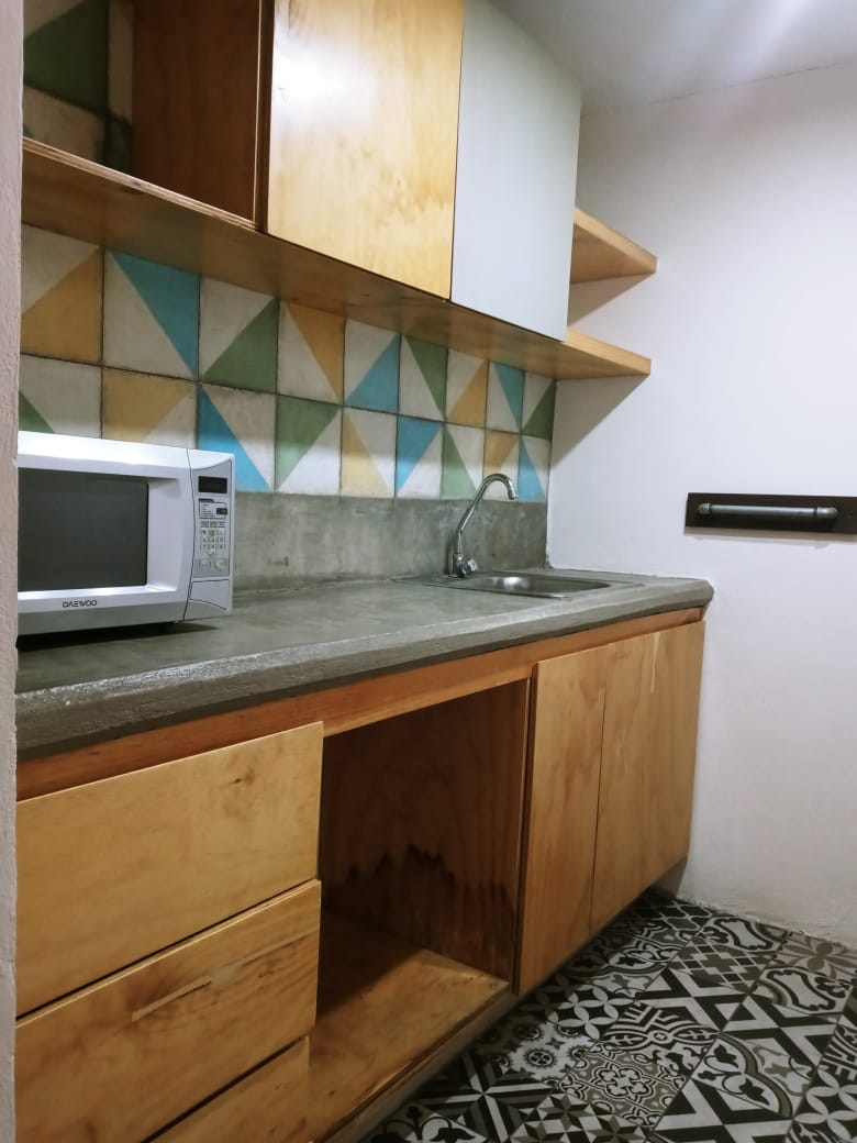 A shared kitchen area is shown with wooden cabinetry and a concrete countertop. Colorful geometric tiles adorn the wall, while a microwave is positioned on the countertop above a sink. The floor features patterned tiles, enhancing the space's overall character.