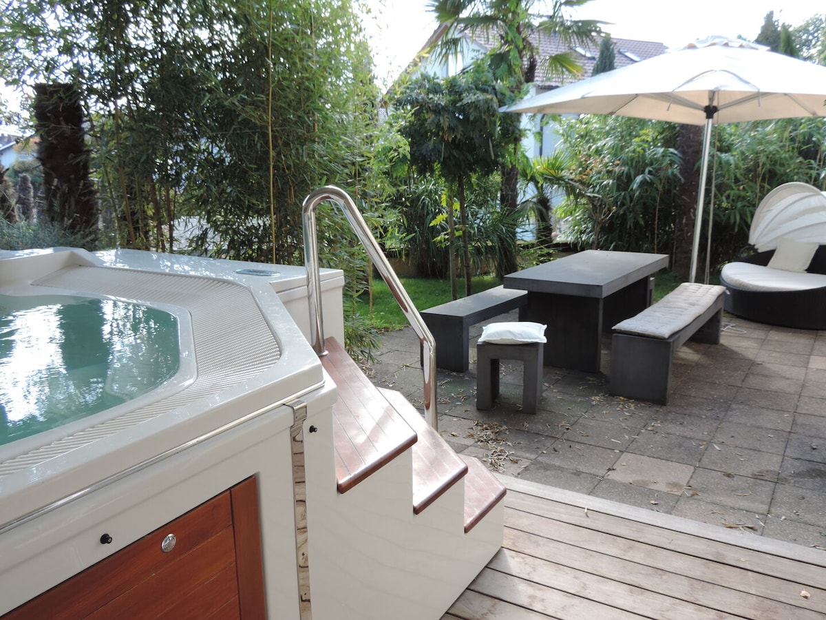 A private garden area features a jacuzzi with sturdy steps leading into it. Nearby, outdoor seating includes a long table with benches and a comfortable lounge chair, all set within a landscaped environment of greenery and palm trees.