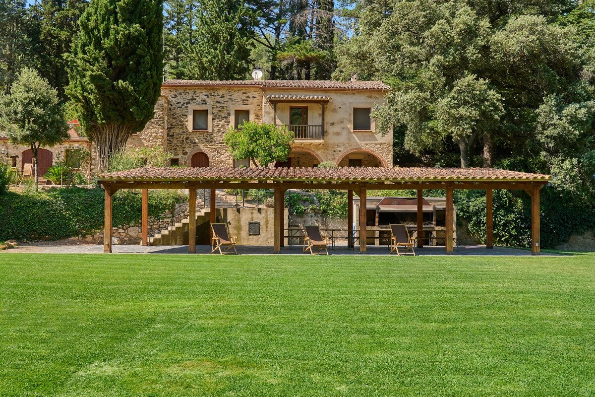 The exterior of the property showcases a spacious lawn framed by the house's rustic stone façade. A covered terrace with wooden beams offers shaded seating, surrounded by lush greenery and trees, creating a peaceful environment for relaxation.
