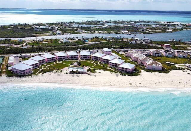 An aerial view captures the beachfront condo complex, displaying several buildings arranged in a circular layout near the shoreline. Palm trees and sandy beach outline the turquoise waters, while nearby islands and serene ocean waves complete the scenic coastal landscape.