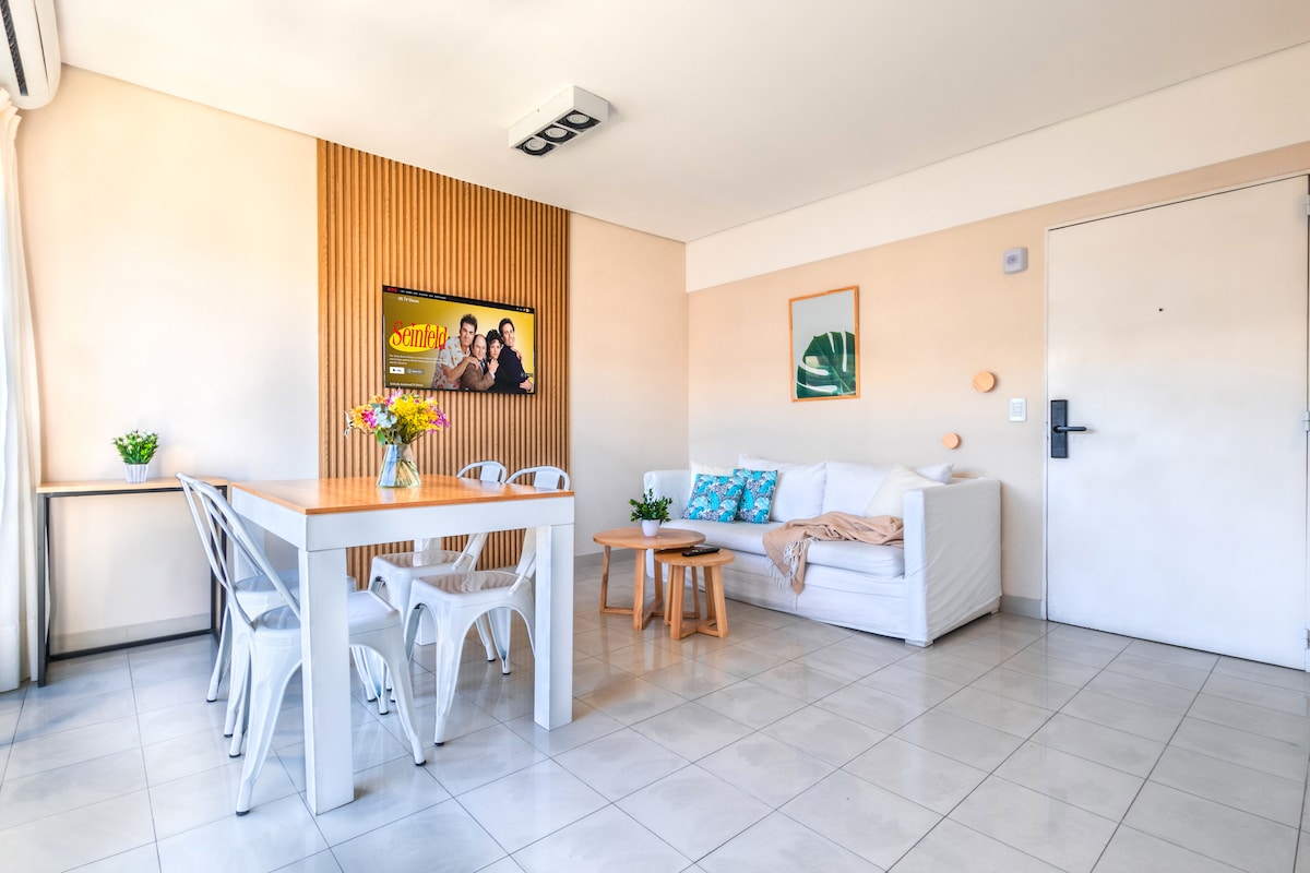 A bright living area is displayed, featuring a white sofa and a small wooden table with a floral centerpiece. A dining table with white chairs stands nearby, and a wall-mounted TV is visible, accompanied by decorative wall art. Natural light enters through the large window.