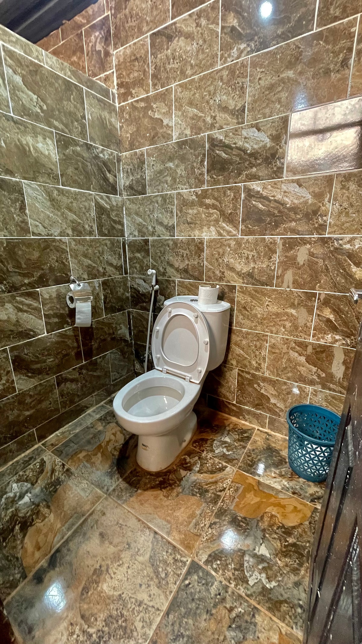 A spacious bathroom is shown, featuring tiled walls with a marble-like pattern and a clean toilet. A small basket and toilet paper are visible on the nearby shelf, providing essential amenities for guests. Neutral tones contribute to a simple yet functional environment.