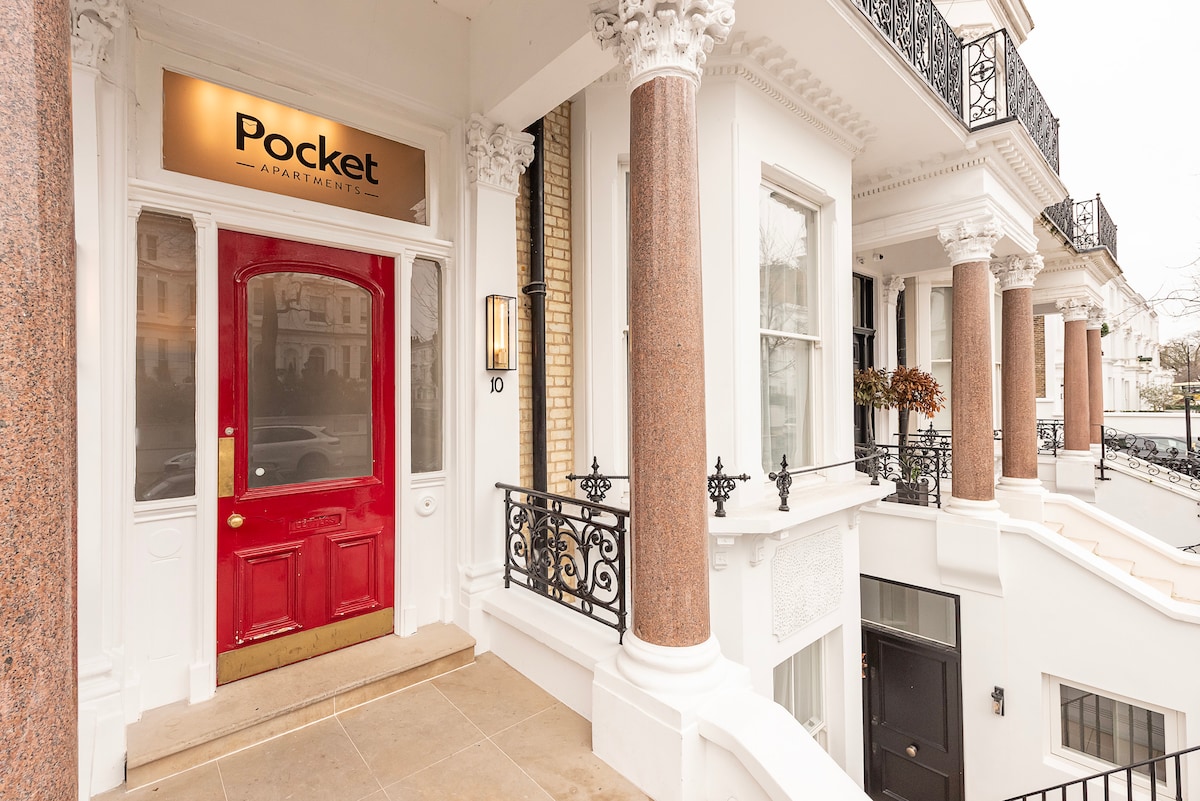 The entrance to the building features a prominent red door framed by decorative stone columns and ornate railings. A stylish sign displays the name 'Pocket Apartments,' enhancing the welcoming aspect of the entrance. The area is well-lit, contributing to a pleasant atmosphere.
