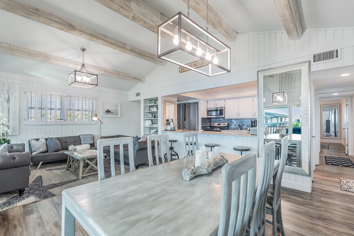 The open living and dining area features a spacious layout with natural light streaming through large windows. A wooden dining table with six chairs is centrally located, while comfortable lounge seating and modern light fixtures create an inviting atmosphere.