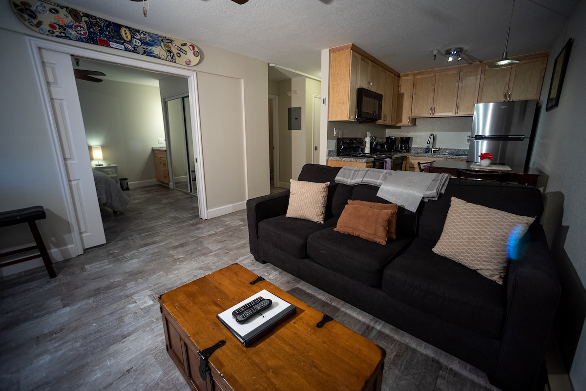 A comfortable living room space is showcased with a dark sofa featuring accent pillows. A wooden coffee table sits in front, with a remote control on top. The kitchen area is visible in the background, and a bedroom doorway is partially seen through pocket dividers.