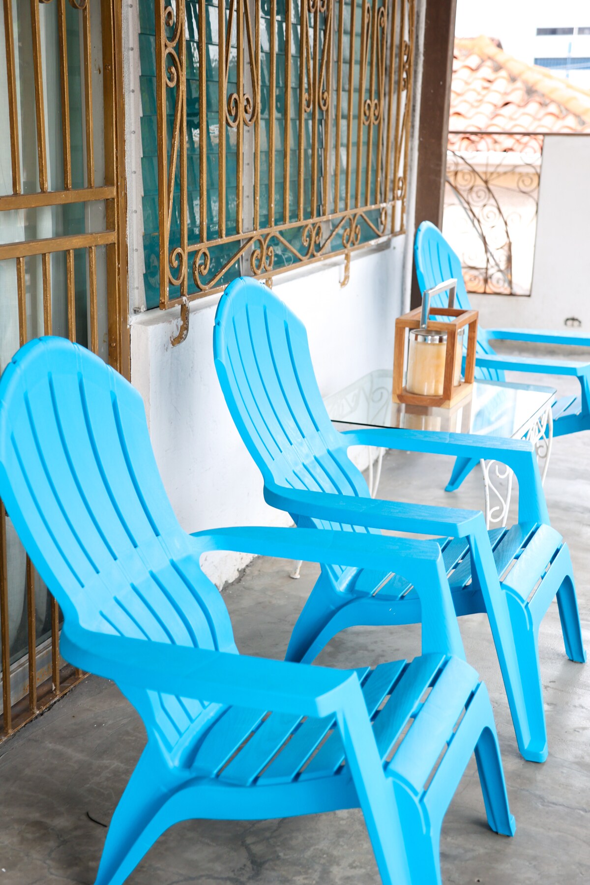 A small outdoor seating area features bright blue chairs arranged around a glass-top table, complemented by a wooden lantern. The space is enclosed with a decorative railing, providing a view of the surroundings.