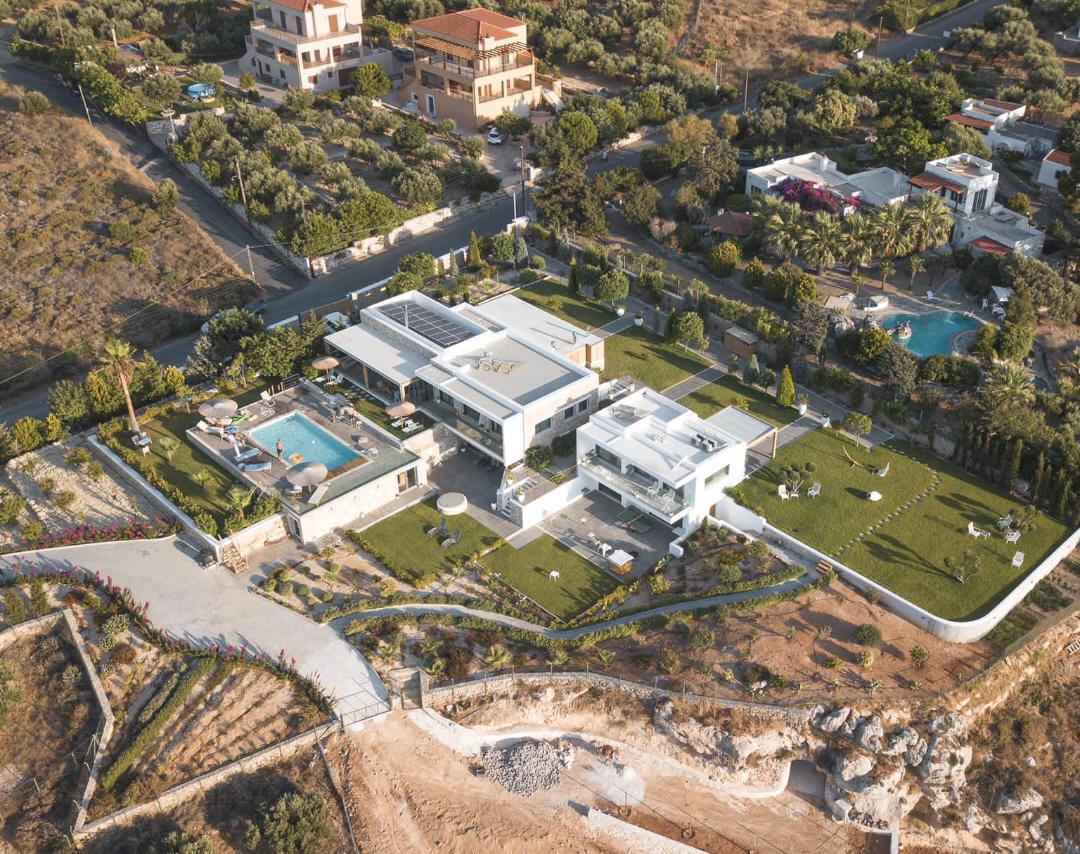 An aerial view of a modern villa showcases its expansive layout, surrounded by lush green lawns and carefully arranged gardens. The 50 sqm heated swimming pool is visible alongside the BBQ area, while the villa's sleek architecture is complemented by the surrounding hillside landscape.