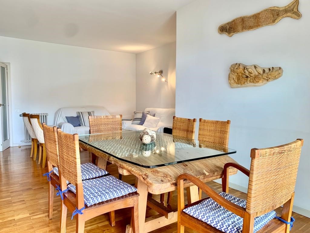 A spacious dining area features a large wooden table surrounded by eight woven chairs. Natural light fills the room, highlighting the minimalist decor, including unique wooden wall art. A comfortable seating area is visible in the background beneath soft white tones.